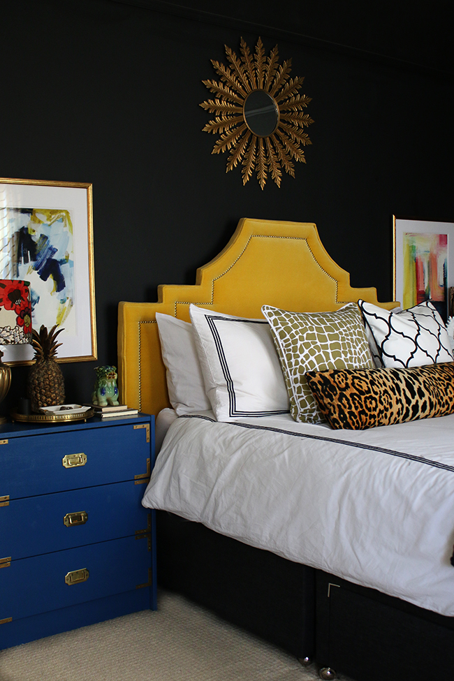 black bedroom with yellow velvet headboard, leopard print cushion, blue rast hack and colourful abstract art - see more on www.swoonworthy.co.uk
