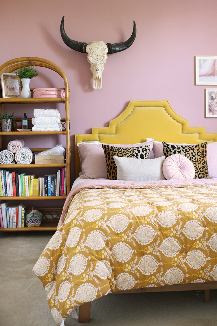 pink bedroom with golden yellow velvet headboard, pink and mustard pattern bedspread and vintage rattan shelving unit